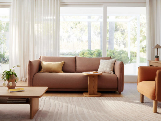 A light pink sofa with a gold cushion, paired with a round wooden table. A potted plant and a beige rug complete the cozy living room setup.
