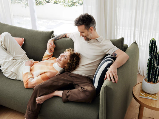 A couple relaxes on a dark green sofa, the woman in a peach and cream sweater, the man in a light beige shirt. A black-and-white striped pillow and a potted cactus are nearby.
