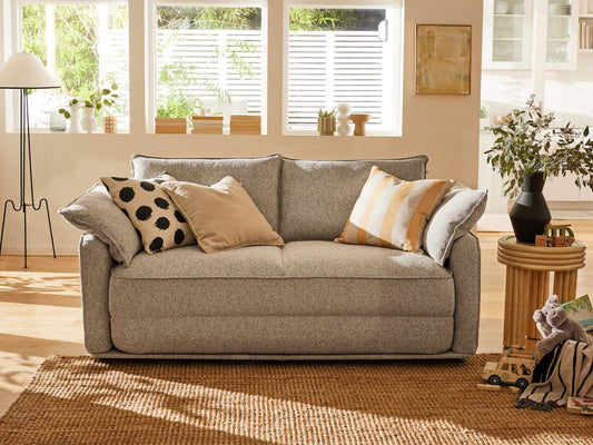 A light grey sofa with patterned and striped cushions sits on a jute rug in a sunlit room with large windows and greenery.