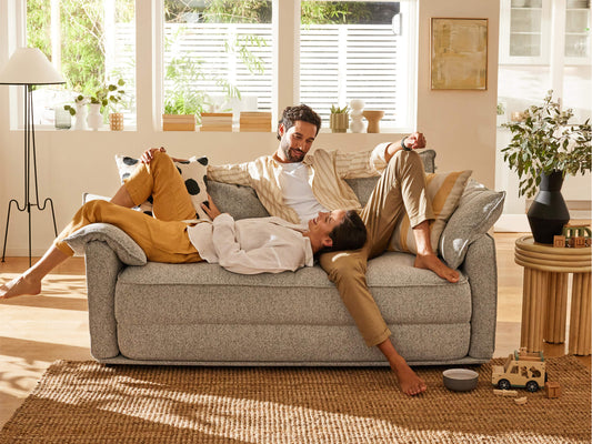 A light grey sofa with a textured surface, two people relax casually; one in tan trousers and a white shirt, the other in mustard pants.