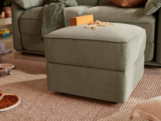 A soft, light green ottoman sits on a beige textured rug, with wooden blocks scattered on top. A dark green throw and a brown pillow rest on a matching sofa in the background.