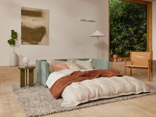 A mint green sofa with white pillows and a rust-colored blanket sits on a textured grey rug, alongside a light wood chair and potted plant.