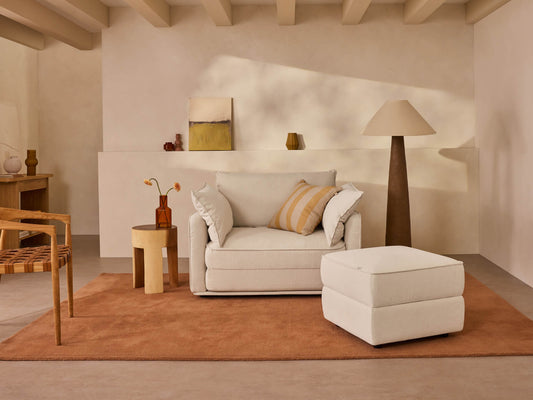 A light beige sofa with striped pillows sits on a rust-colored rug, beside a brown lamp and wooden side table in a softly lit room.