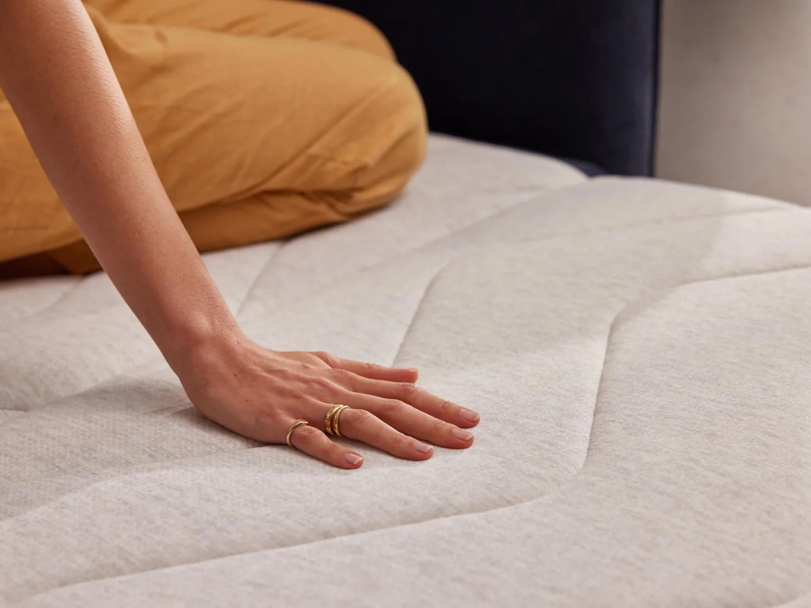 A close-up of a hand resting on a light grey quilted mattress, with a background of soft golden-colored bedding.