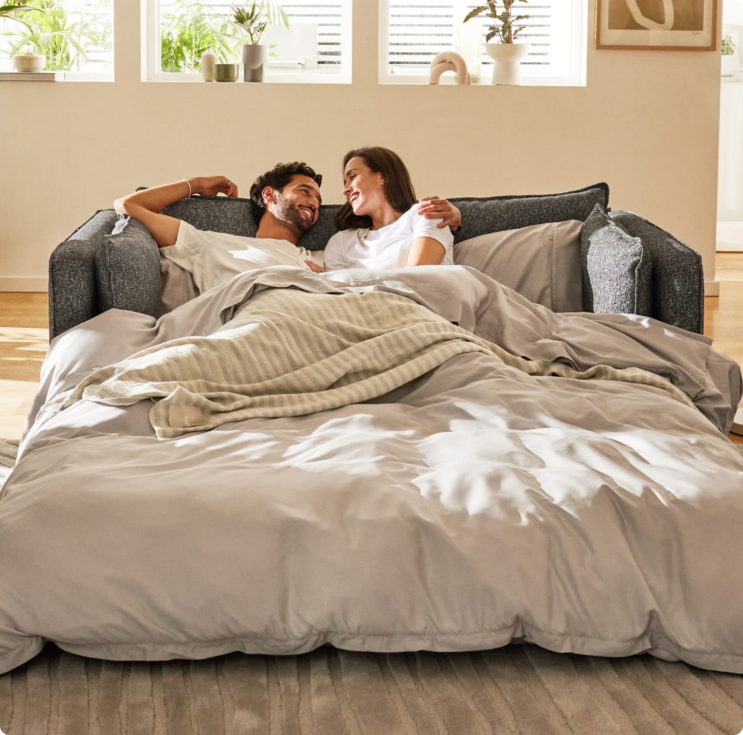 A couple relaxes on a dark grey sofa, covered with a light grey duvet and a patterned cream blanket, smiling and gazing at each other.