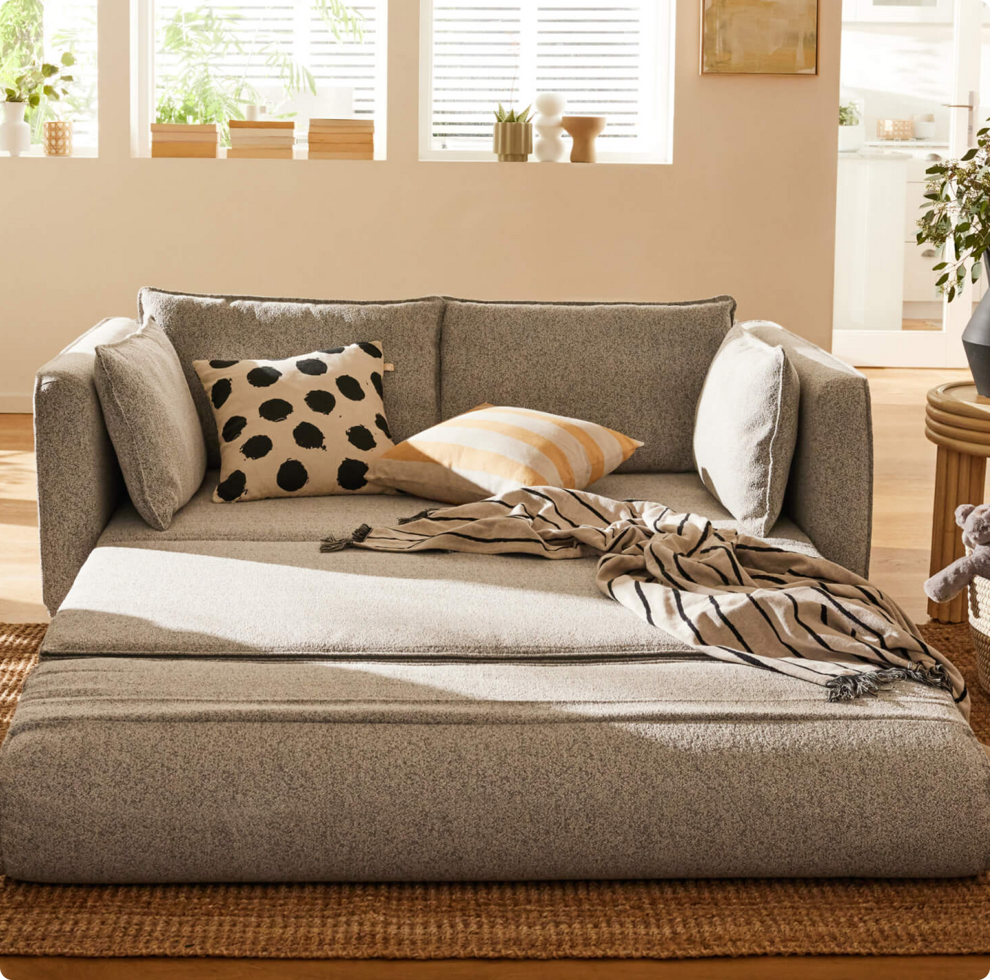 A light grey sofa with black polka dot and yellow striped pillows, draped with a beige striped throw, on a woven rug.