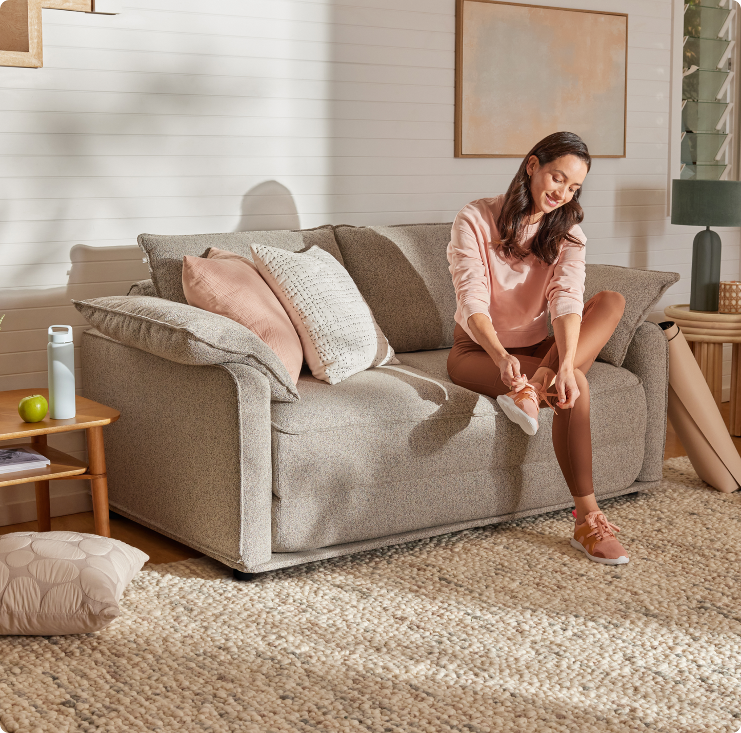 A woman in a light pink sweatshirt and brown leggings sits on a textured light grey sofa, tying her white sneakers. A green apple rests on a wooden table nearby.