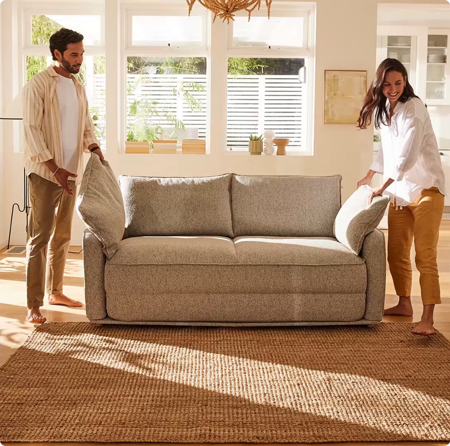 A light grey sofa with large cushions is being arranged by a man in a cream shirt and a woman in a white shirt, barefoot on a tan rug.