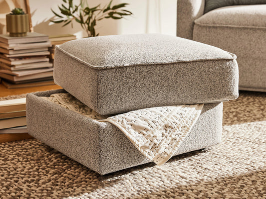 A textured light grey ottoman with a removable top cushion, revealing a soft beige quilt underneath, sits on a woven rug.