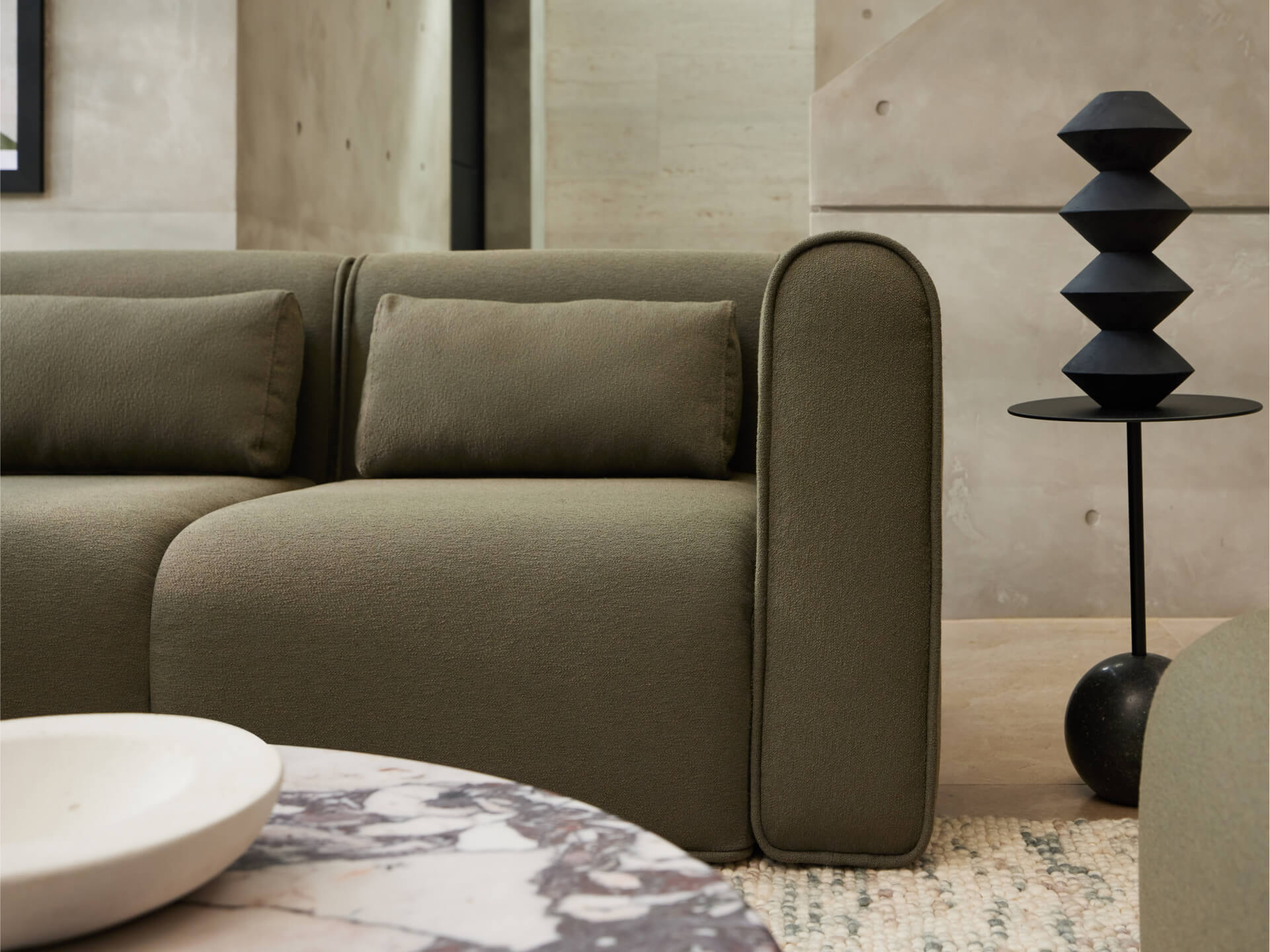 Elegant olive-green fabric sofa with plush cushions, rounded armrest; set in a modern concrete-finish room with a marble coffee table and geometric decor.