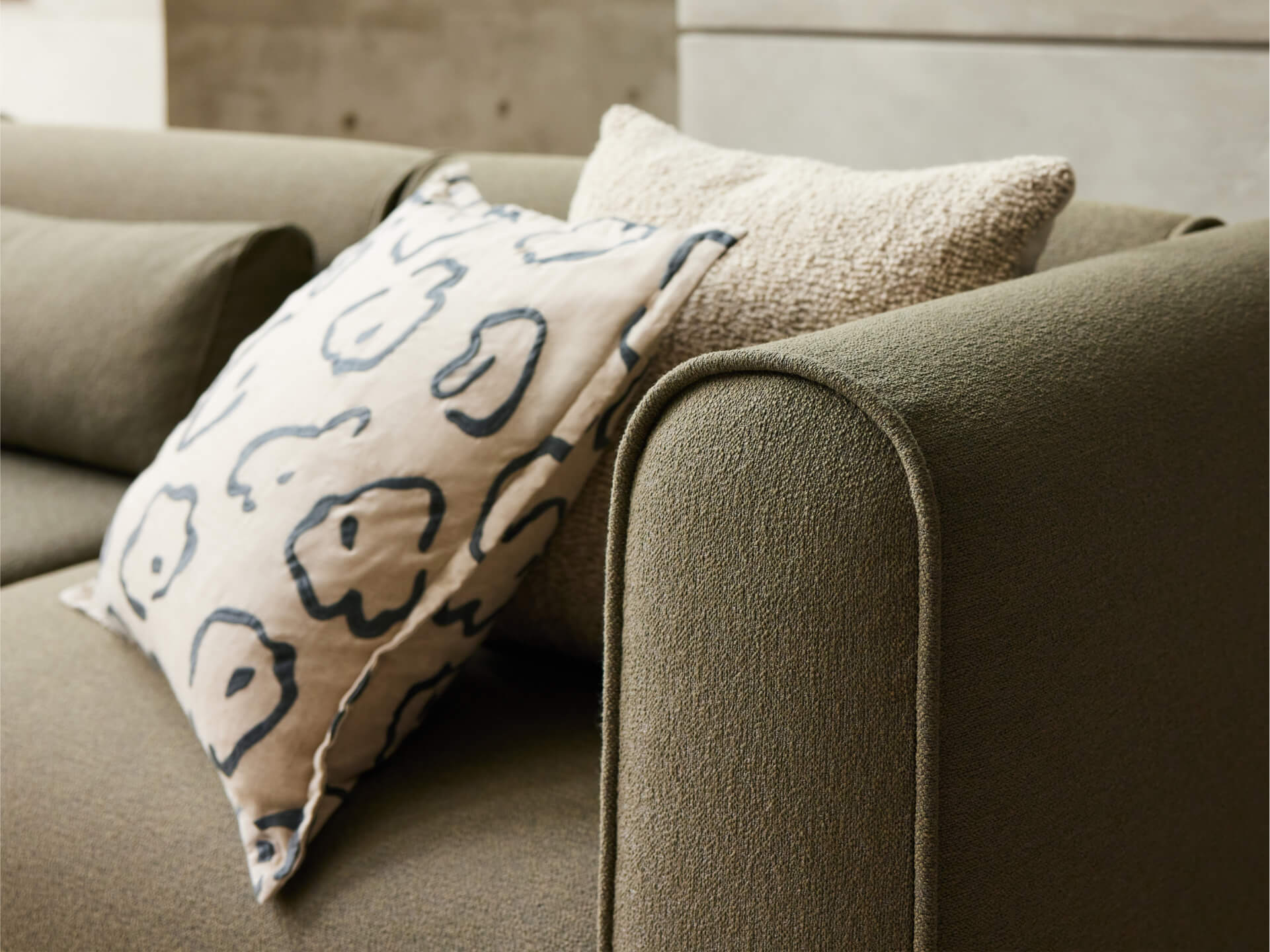 Olive green upholstered sofa with curved armrests, featuring textured fabric. Accented by beige and patterned cushions, set in a modern living space.
