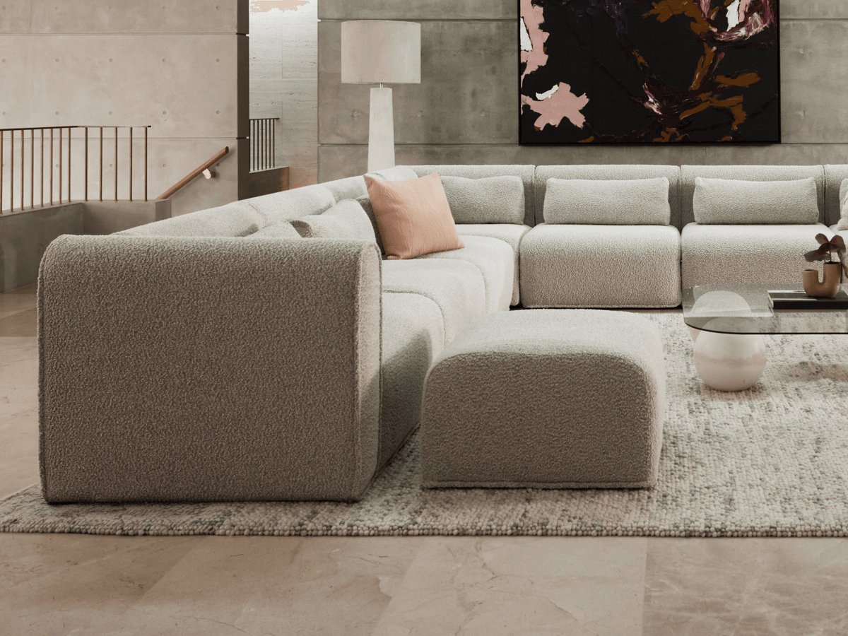 Luxurious L-shaped chaise lounge in soft grey boucle, featuring plush cushions and a matching ottoman. Set in minimalist concrete interior with abstract art.