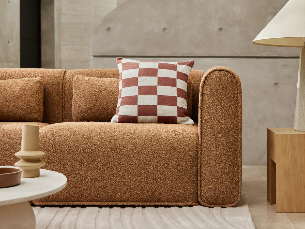 Plush boucle sofa in warm caramel tones with rounded arms and checkerboard cushion, set in a minimalist living room with neutral accents.