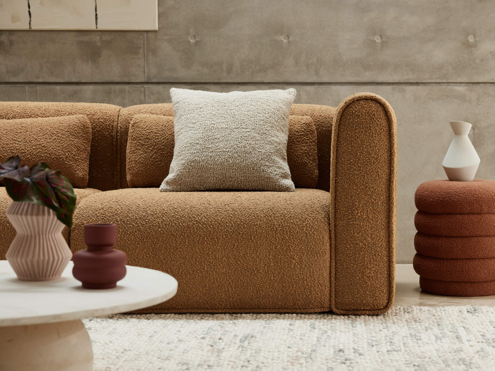 Luxurious boucle sofa in warm caramel hue, featuring plush cushioning and a curved silhouette. Set against a concrete wall, paired with chic decor elements.