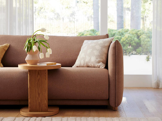 A soft, light brown sofa with a thick cushion and a beige floral pillow. Beside it is a round wooden table holding a pink vase with white tulips.