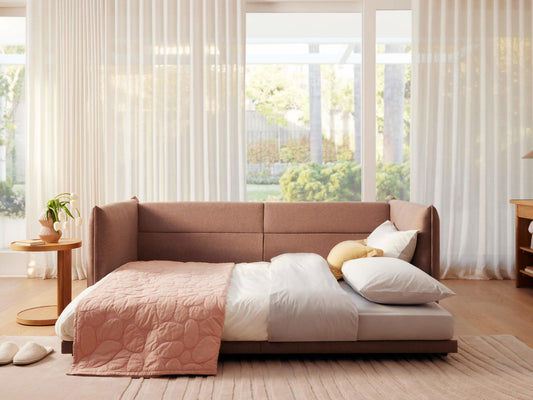 A brown, upholstered sofa partially folded down as a bed, with white sheets, a pink quilt, and decorative pillows in white and yellow. Sunlight streams through sheer white curtains.