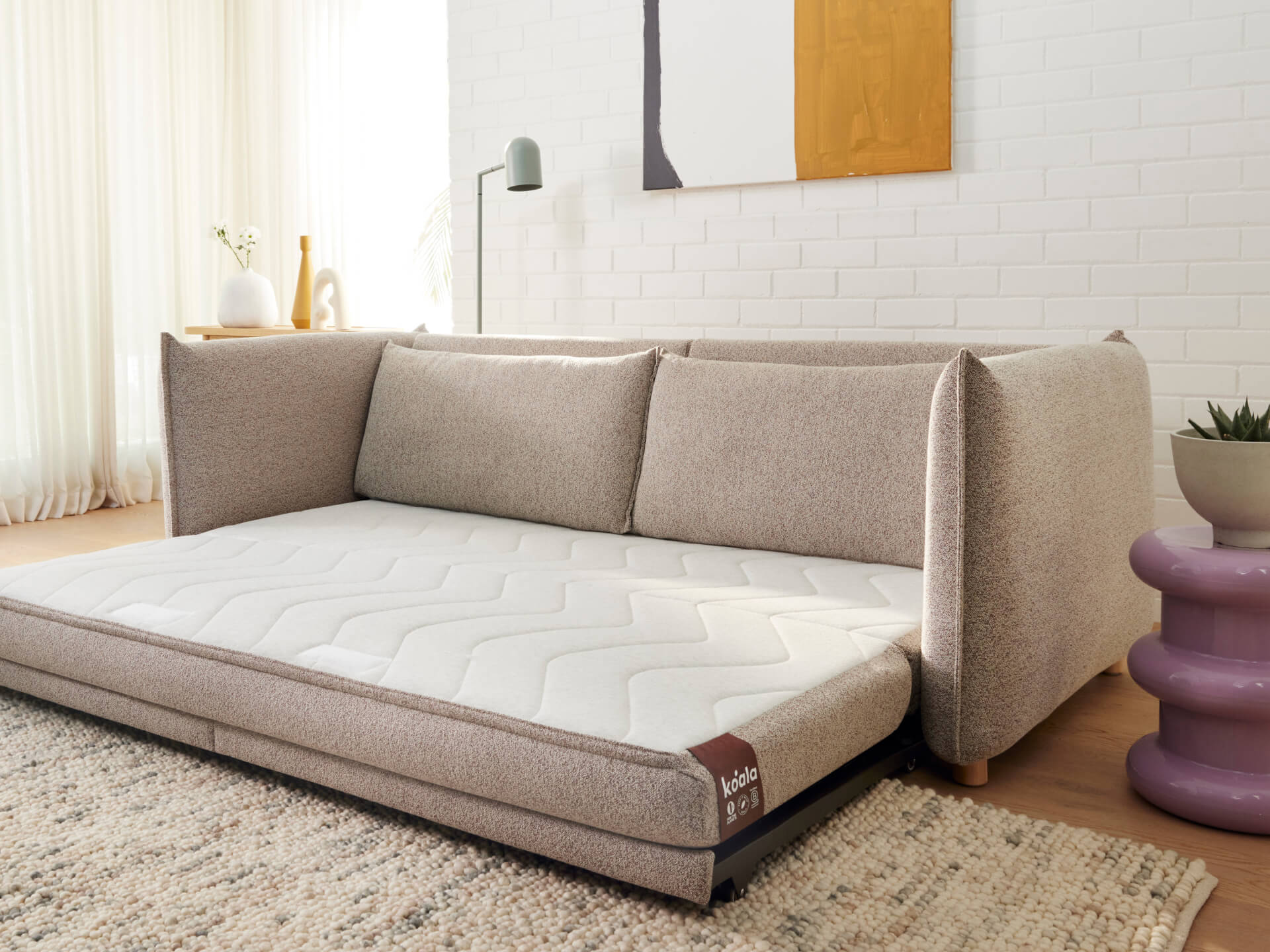 A light brown sofa with plush cushions, featuring a white, wavy-patterned mattress. Beside it is a purple side table and a textured beige rug.