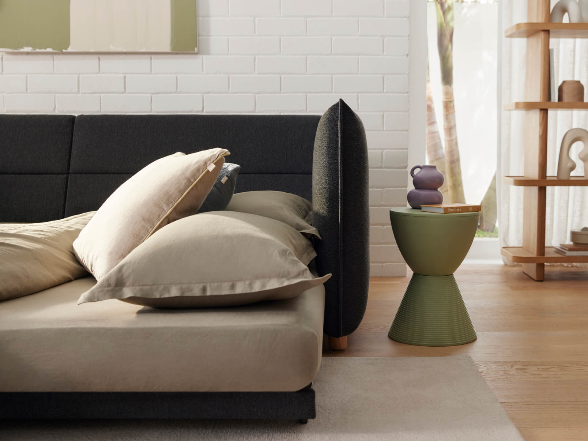 A dark grey bed with beige and green pillows, beside a green ribbed side table holding a purple vase and a book, in a bright room.