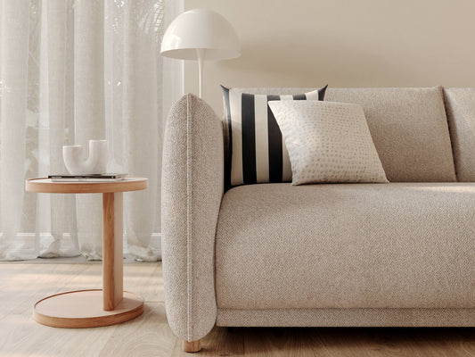 A light beige sofa with a curved armrest, a patterned cushion, a black and white striped pillow, and a round wooden side table with a white lamp.