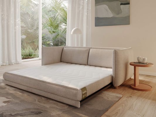 A light grey sofa bed with a white mattress sits in a bright room with large windows and a wooden floor, accompanied by a small orange side table.