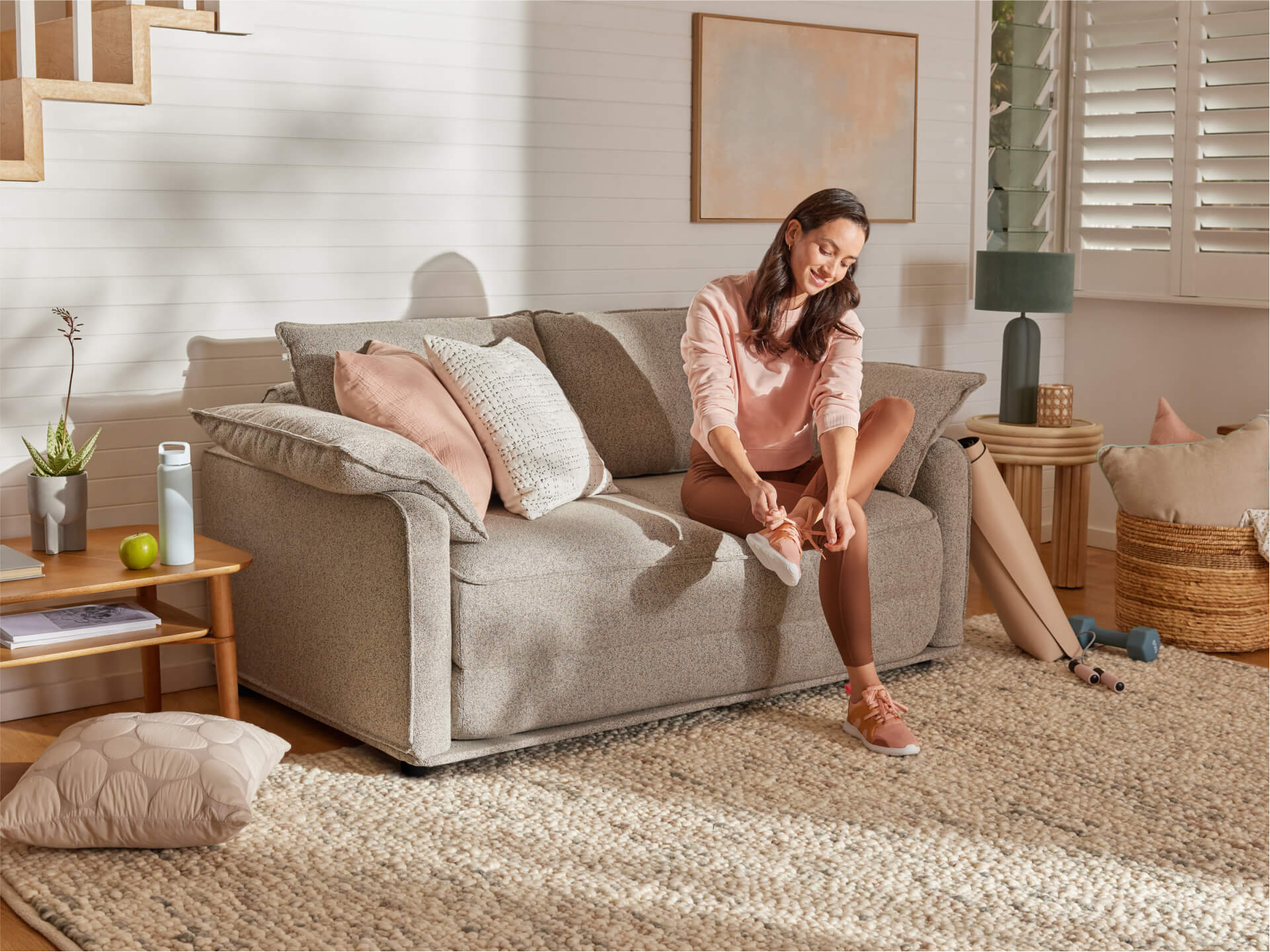 A woman in a light pink sweater sits on a light grey sofa, tying her sneakers. The room has warm tones and natural light, with a beige rug and houseplants.