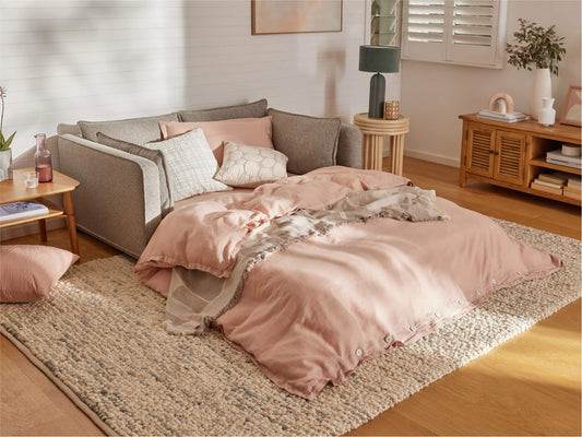 A cozy scene featuring a light grey sofa with soft pink bedding and pillows, a woven rug, and wooden furniture in a warmly lit room.