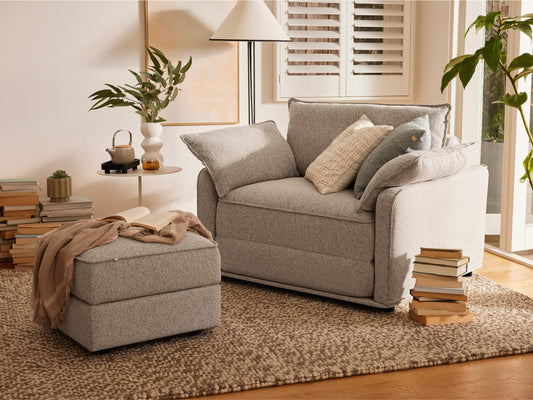 A light grey armchair with two cushions sits on a textured beige rug beside a small side table and a potted plant, in a bright room.