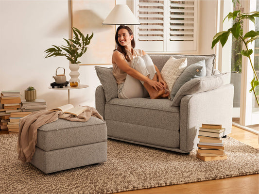 A woman in a light grey sofa smiles while sitting with her legs crossed. A tan throw and small grey ottoman are nearby on a textured beige rug.