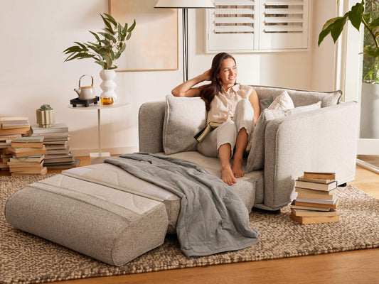 A woman in a light beige top relaxes on a light grey sectional sofa with a light grey throw. Books are stacked nearby on a textured, beige rug.