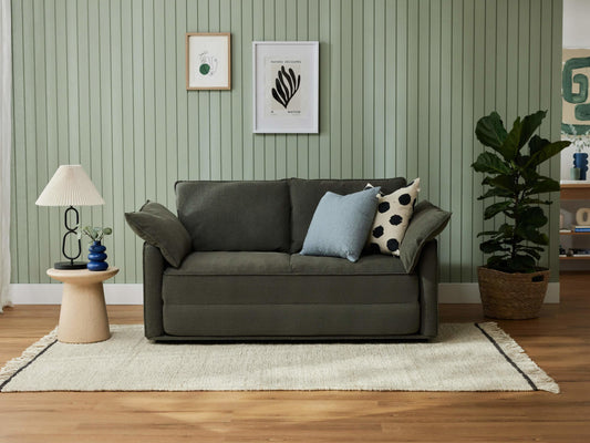 A dark green sofa with two pillows, one light blue and one with black dots, beside a beige table lamp and a potted plant against a light green wall.