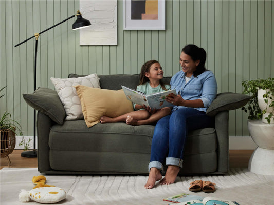 A woman and girl sit on a dark green sofa, sharing a book. The room features light green walls, a black lamp, and a white decorative plant pot.