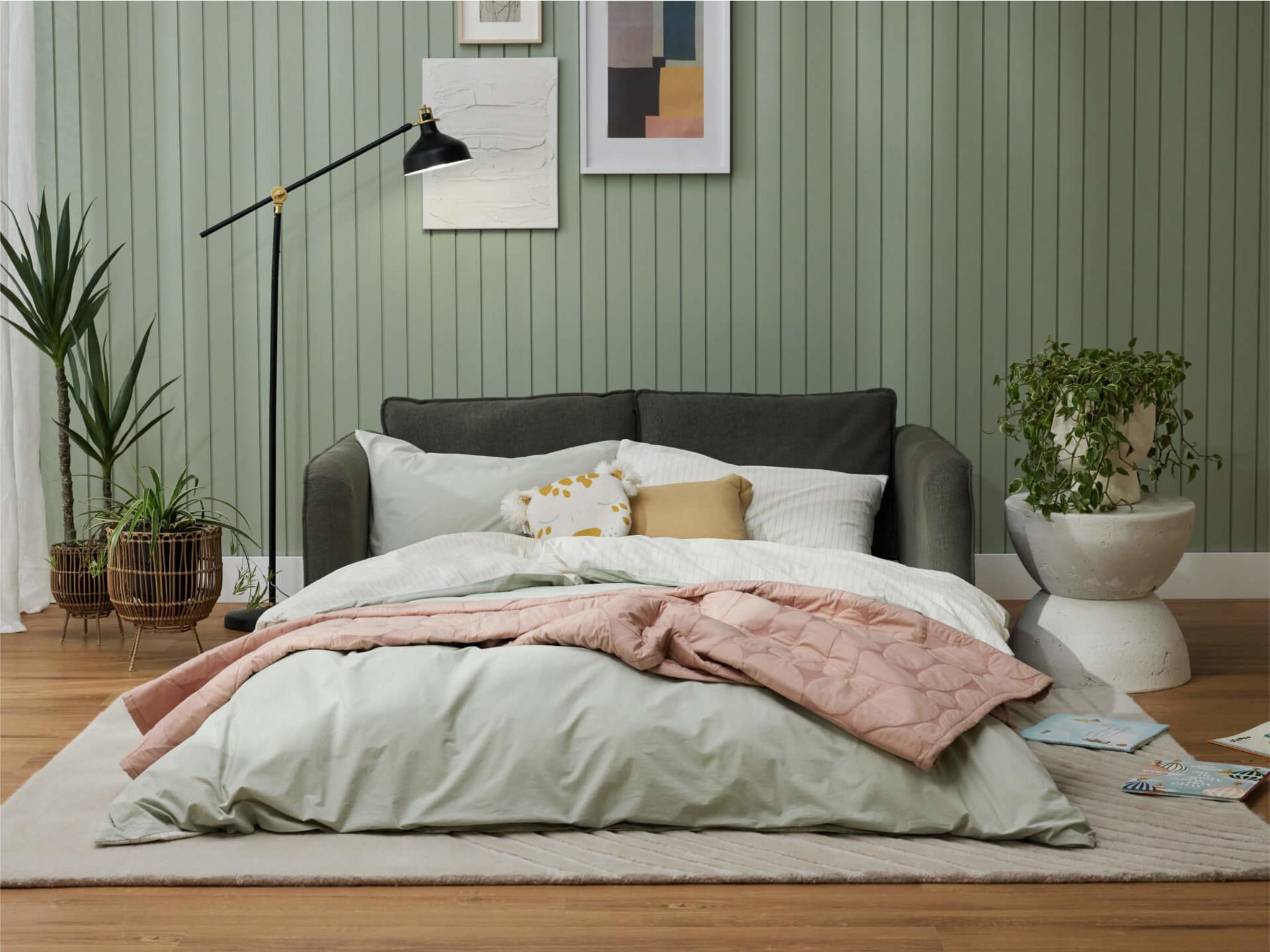 Cozy bedroom with light green walls, a dark grey sofa, pastel pink and mint bedding, a black floor lamp, and plants in woven pots.