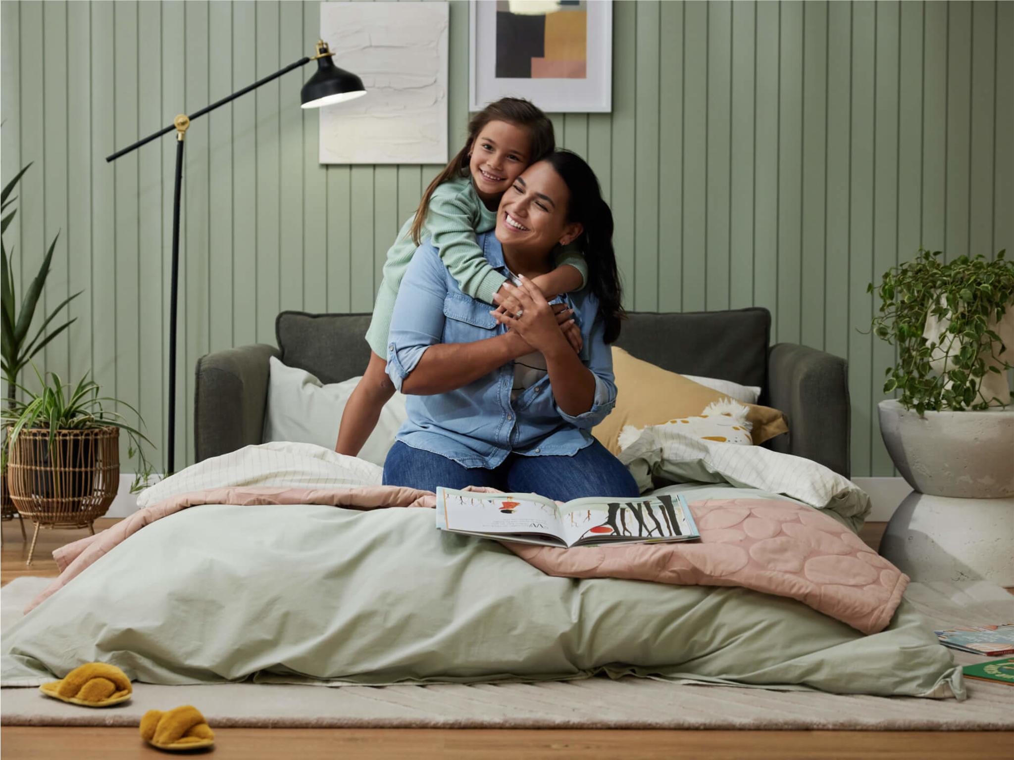 A woman sits on a soft green blanket with a child hugging her. They smile, surrounded by a light green wall, plants, and a brown sofa.