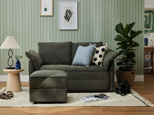 A dark green sofa with patterned pillows, a light beige lamp, and a brown side table. A woven pot plant and magazine on a light rug.