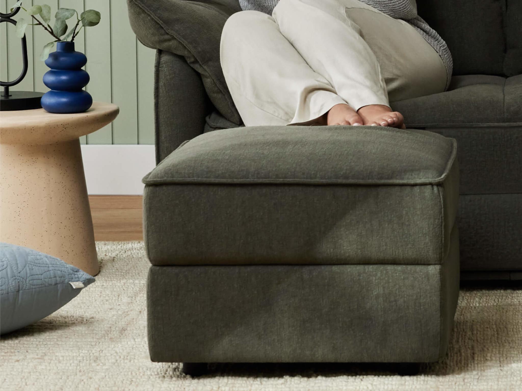 A green ottoman sits on a light beige rug, near a light tan table with a blue vase and a plant, while a person rests feet on the sofa.