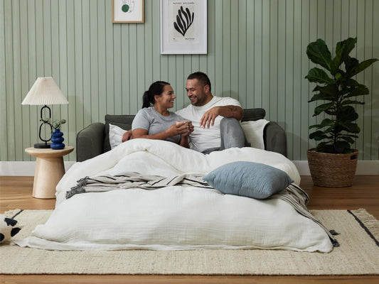 A smiling couple sits on a dark grey sofa with a white blanket. Pale green walls and a potted plant complete the cozy room. A lamp and side table add decor.