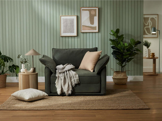 A dark green sofa with light pillows sits on a jute rug, near a round wooden table, lamp, and potted plants against a green wall.