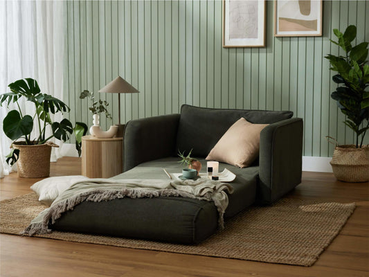 A dark green sofa with a beige pillow, draped with a light grey throw, beside a round wooden table and potted plants on a woven rug.