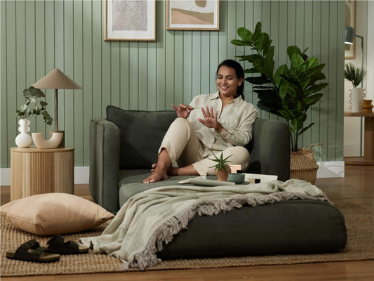 A woman in a light beige shirt sits on a dark green sofa, surrounded by plants, a cream lamp, and a soft pink pillow on a woven rug.