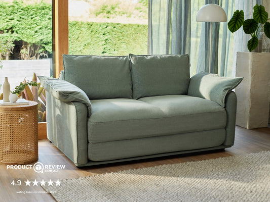 A light green sofa with oversized cushions sits beside a rattan side table and a textured rug, illuminated by natural light through sheer curtains.