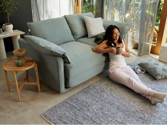 A woman in a white tank top sits on a light grey sofa, holding a green mug. She wears light pink sweatpants, with a soft grey rug below.