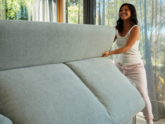 A woman in a white tank top and light pink sweatpants joyfully lifts a light grey sofa cushion in a bright room with sheer curtains.