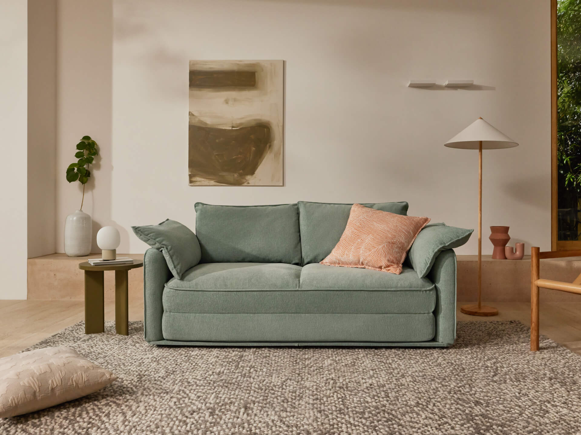 A cozy mint green sofa with a coral pillow sits on a patterned grey rug in a light, airy room with natural elements and decor.