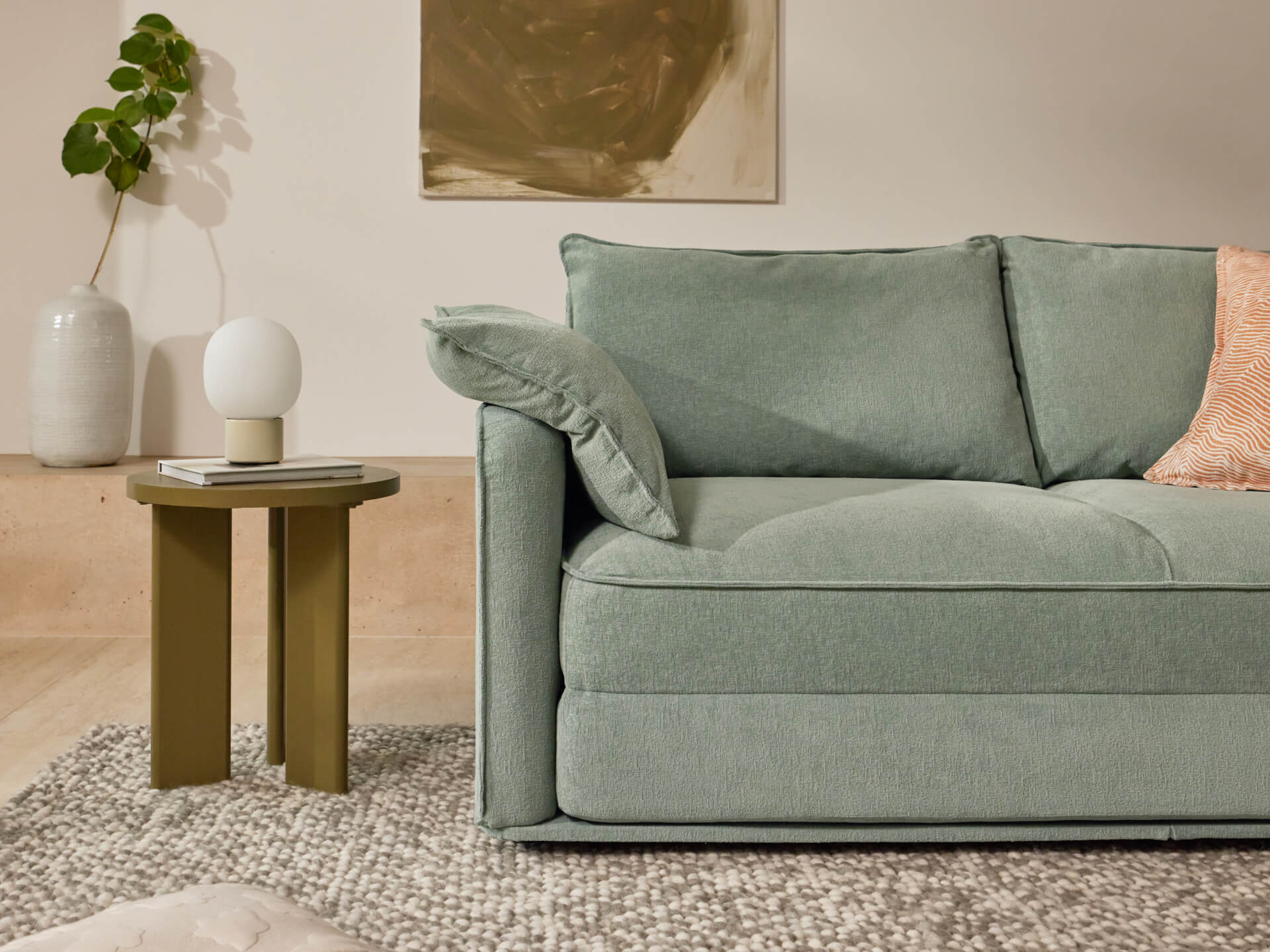 A light green sofa with a loose cushion, a round white lamp on a dark olive green side table, and a patterned grey rug, all in a neutral room.