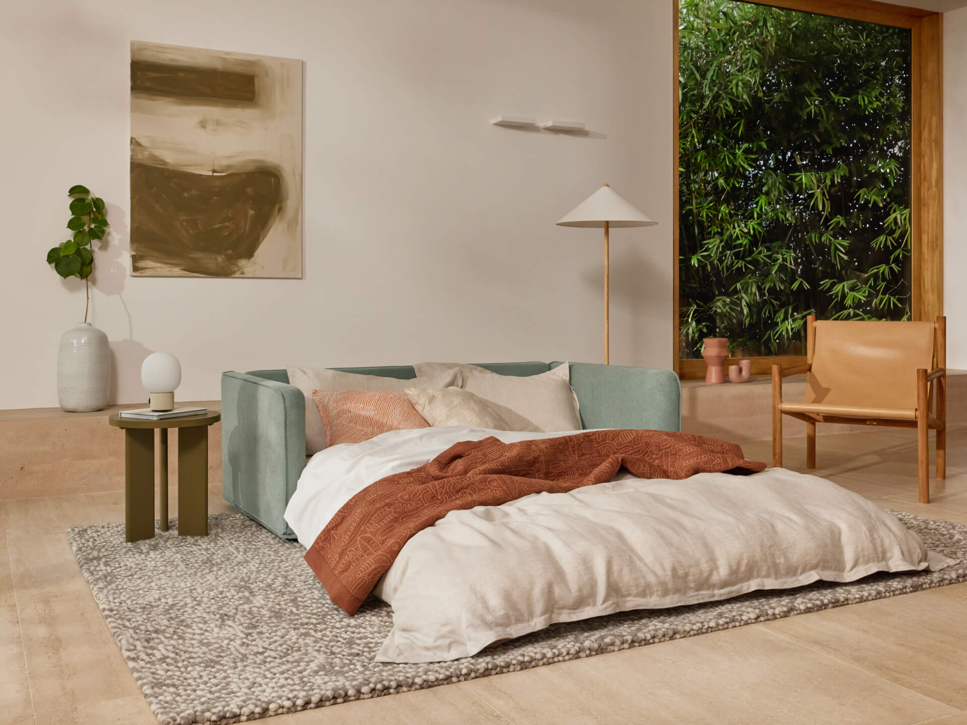 A mint green sofa with white pillows and a rust-colored blanket sits on a textured grey rug, alongside a light wood chair and potted plant.