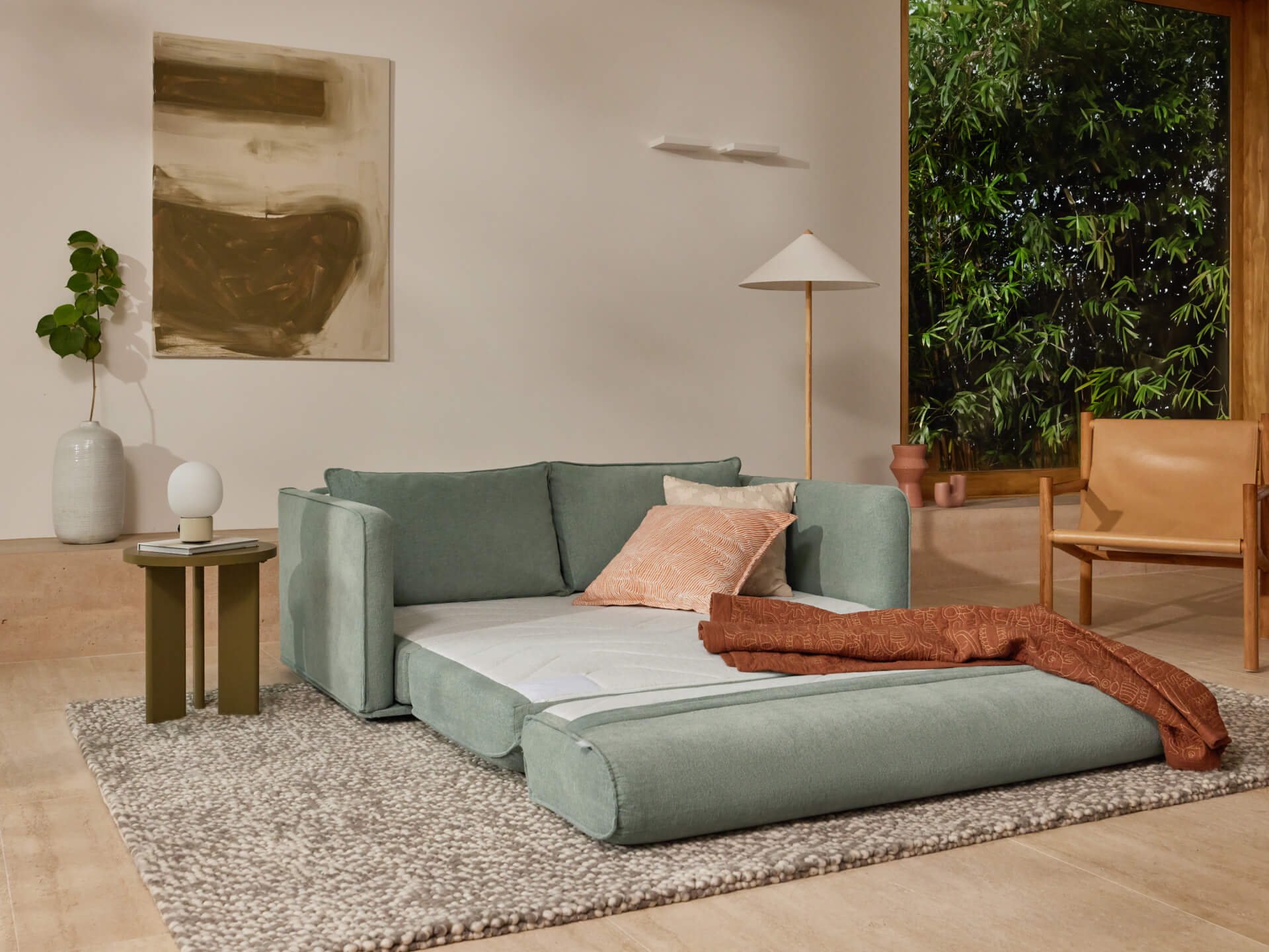 A light teal sofa with a peach pillow and a rust-colored throw, paired with a white lamp and greenery in a bright, airy room.