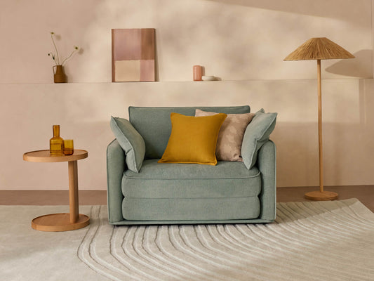 A soft, light teal armchair with yellow and beige pillows next to a round wooden table and a tall, beige lamp on a cream rug.