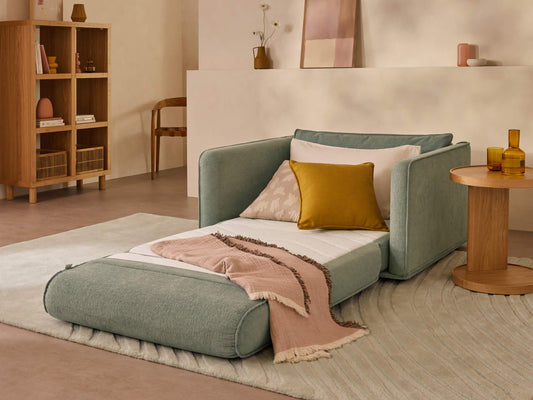 A mint green chaise lounge with a white cushion, yellow pillow, and pink throw blanket sits on a light beige rug in a soft-toned room.