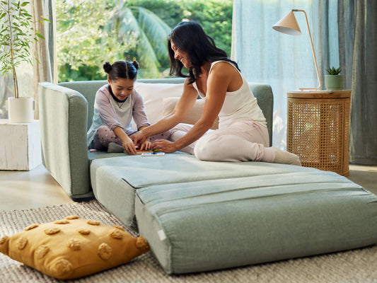 A light green sofa with a low back, a girl in grey pajamas, and a woman in white top and pink pants play together on the floor. A yellow textured pillow and a woven side table are nearby.