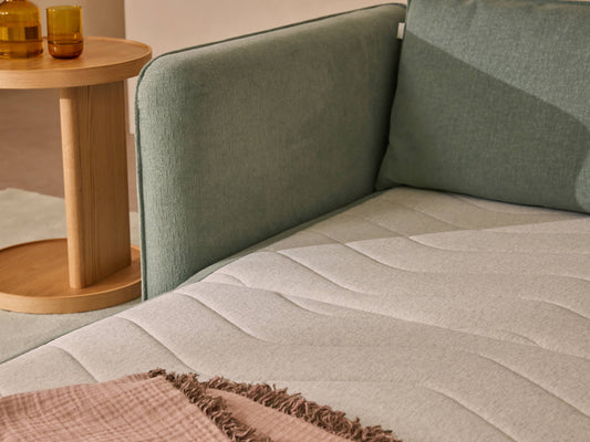 A light grey mattress rests against a mint green sofa arm, alongside a wooden side table with amber glasses and a soft pink throw blanket.